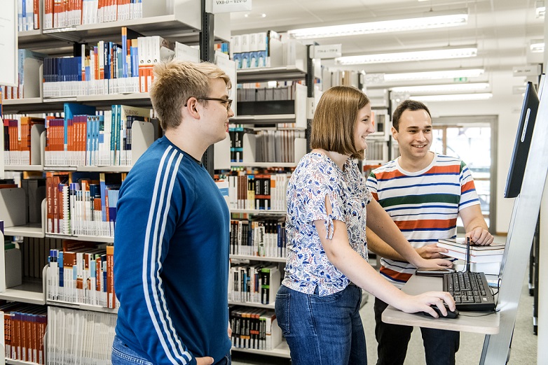 Zwei Studenten und eine Studentin stehen vor einem festinstallierten Computer, auf den sie lächelnd drauf schauen.