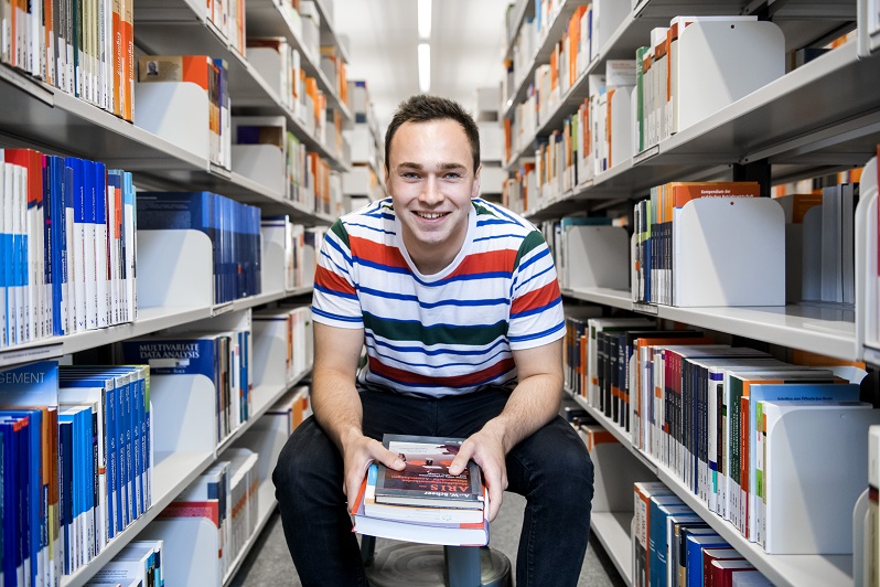 Ein Student sitzt auf einem Hocker zwischen zwei Bücherreihen in der Bibliothek am Campus Lingen.