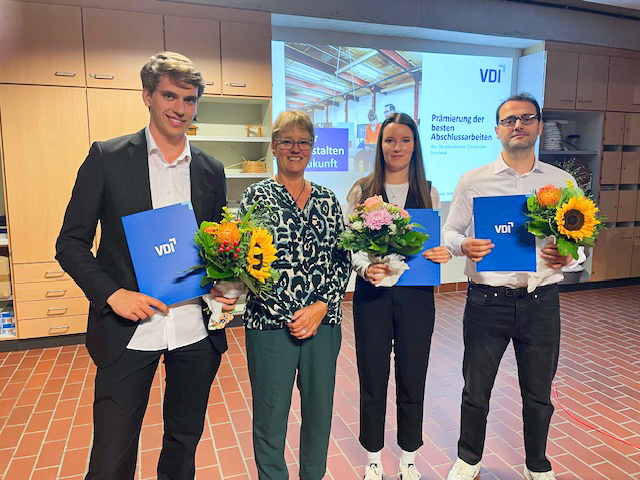 Drei Studierende halten Urkunden fest und lächeln in die Kamera. In der Mitte steht ihre Professorin.