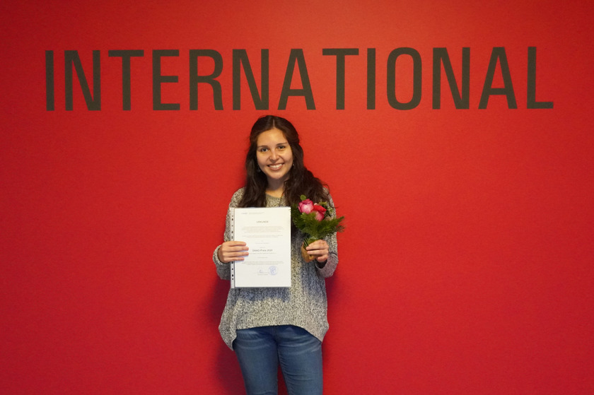 Lorena Escobar Arispe steht vor einer roten Wand mit dem Schriftzug International und hält eine Urkunde und einen Blumenstrauß in der Hand