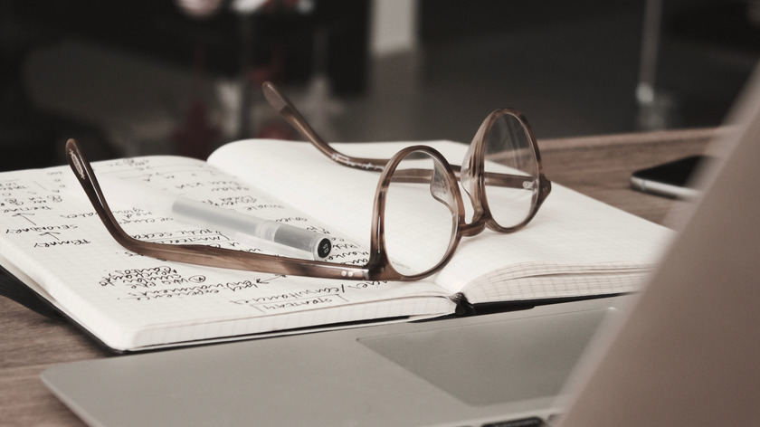 Auf einem Tisch liegt ein beschriebenes Notizbuch, auf dem ein Stift und eine Brille liegen. Daneben liegt ein Smartphone und davor steht ein Laptop.