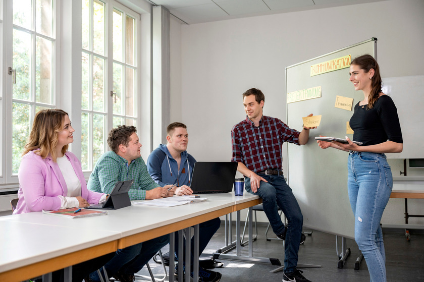 Drei Studierende sitzen an einem Tisch, zwei Studierende stehen in einem Vorlesungsraum und unterhalten sich