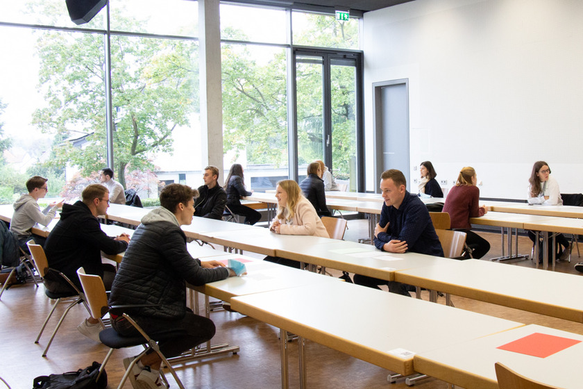 Mehrere Studierende zietzen in einem lichtdurchfluteten Raum. Sie sitzen sich immer zu zwei gegenüber. Zwischen ihnen stehen zwei Tische. Sie unterhalten sich. 