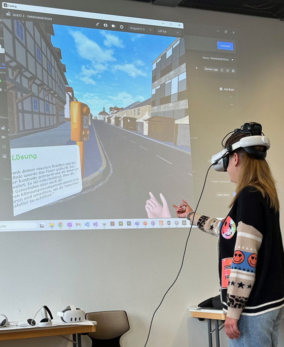 Eine Person mit VR-Brille interagiert mit einer virtuellen Umgebung, die eine Stadt mit Gebäuden und Menschen zeigt. Die Person zeigt mit der Hand auf einen Bildschirm, der die VR-Anwendung steuert. Das Bild zeigt einen Prototyp des Forschungsprojekts KlimKoVR, das Studierenden helfen soll, die Auswirkungen des Klimawandels zu verstehen.