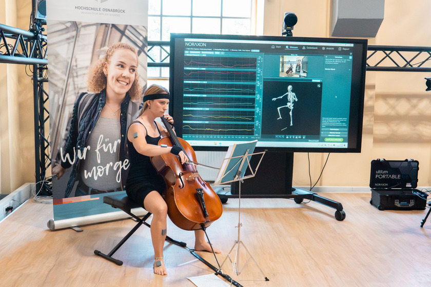 auf dem Bild ist eine Cellospielerin zu sehen in einem schwarzen Kleid und barfuß. An den Beinen und Armen sind Elektroden befestigt. Rechts dahinter steht ein großer Monitor, der ihre Bewegungen aufzeichnet.