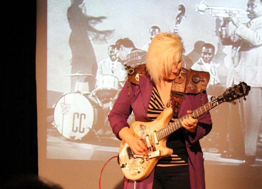 Auf dem Bild sieht man eine blonde Frau vor einer Leinwand, die E-Gitarre spielt.
