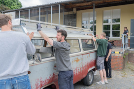 Mehrere Personen heben gemeinsam ein großes, rechteckiges Objekt auf das Dach eines alten, rot-weißen VW-Busses