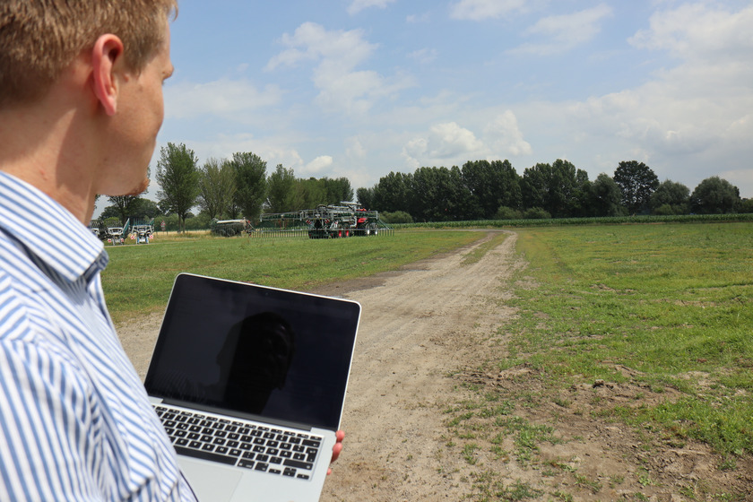 Mann hält Laptop in der Hand und schaut auf ein Feld, über das ein Traktor fährt.