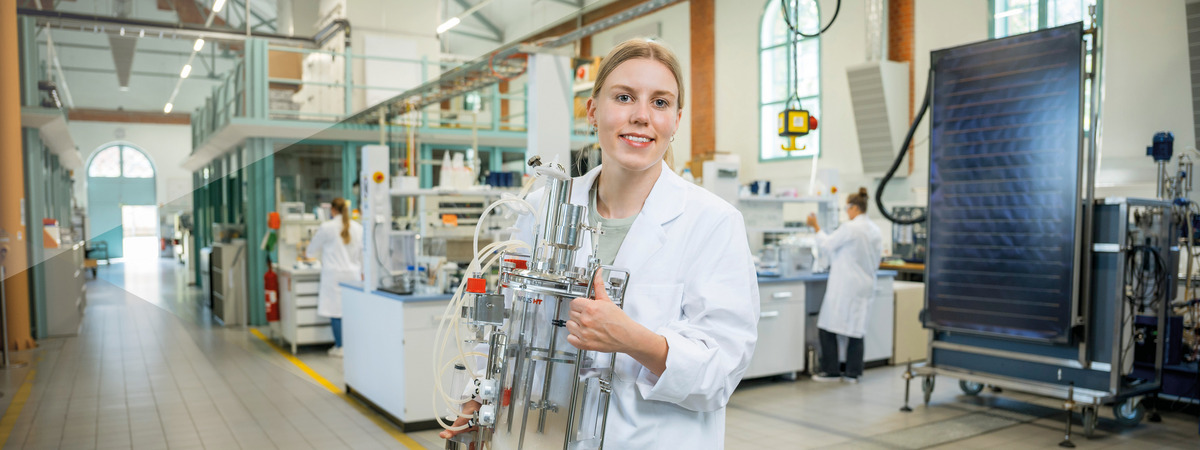 Wissenschaftlerin im Laborkittel hält einen technischen Glasreaktor in einem Labor.