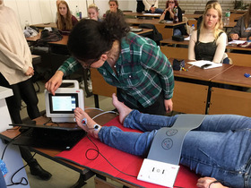 Studentinnen sitzen in einem Hörsaal und sehen zu, wie eine Studentin das BIA-Messgerät an einer auf einem Tisch liegenden Studentin ausprobiert.