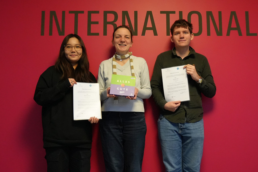 Drei Personen stehen vor einem kräftig pinken Hintergrund mit dem großen, schwarzen Schriftzug "INTERNATIONAL". Sie halten Zertifikate und ein Geschenk und lächeln in die Kamera. Die linke Person ist eine junge Frau mit langen, dunklen Haaren. Sie trägt eine schwarze Kapuzenjacke und eine Brille. Sie hält ein Zertifikat mit beiden Händen und lächelt. Die mittlere Person ist eine Frau mit kurzen, blonden Haaren und trägt einen hellgrauen Pullover und einen gestreiften Schal. Sie hält ein grün-lila Geschenkpaket mit der Aufschrift "ALLES GUTE" und steht zwischen den beiden anderen Personen. Die rechte Person ist ein junger Mann mit kurzen, braunen Haaren. Er trägt ein dunkelgrünes Hemd und Jeans. Er hält ein Zertifikat in seiner rechten Hand und zeigt darauf mit seiner linken Hand. Alle drei stehen entspannt und glücklich nebeneinander, und der Hintergrund betont den internationalen Kontext des Bildes.