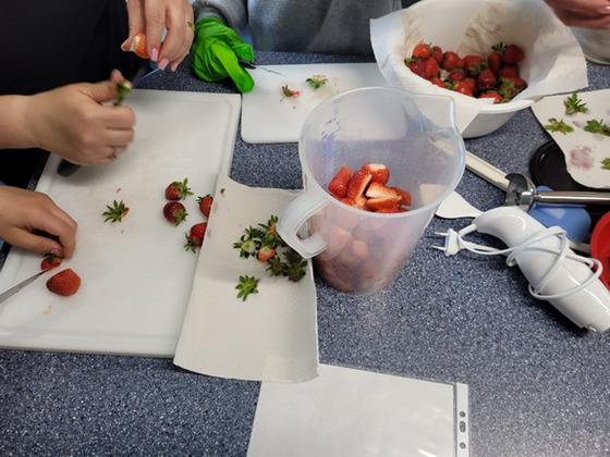 Das Bild zeigt Hände, die Erdbeergrün abschneiden und Erdbeeren und Grün in Schalen verteilen.