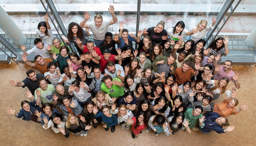 Eine Gruppe junger Studierender wird von oben fotografiert. Sie sehen fröhlich aus, lachen und winken in die Kamera. 