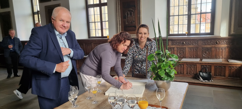 Drei Personen stehen vor einem Tisch, auf dem das Osnabrücker Gästebuch liegt. Links der Herr steht aufrecht und schaut in die Kamera, die Dame links daneben schreibt ins Gästebuch und die dritte Person steht recht daneben und schaut ebenfalls in die Kamera.