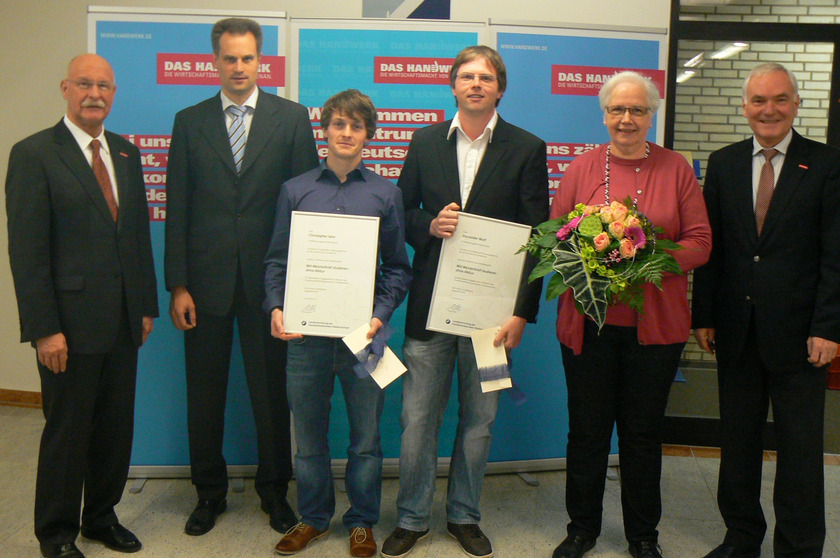 Auf dem Bild sieht man zwei Männer, die Urkunden in der Hand halten. Um sie herum stehen drei Männder und eine Frau.
