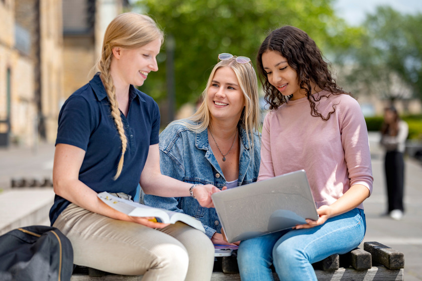 Drei june Frauen sitzen auf draußen auf einer Bank und unterhalten sich. Eine hat einen Laptop auf dem Schoß.