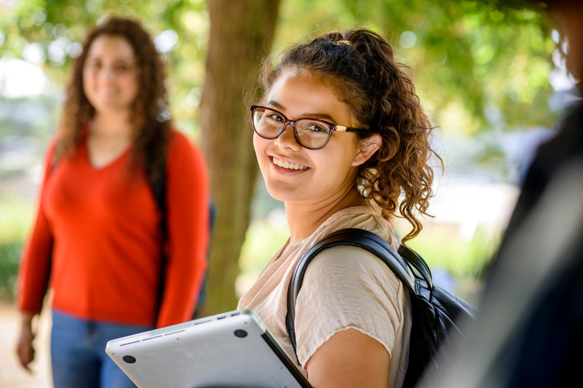 Internationale Studentin, mit brauner Brille, steht mit ihrem Laptop in der Hand unter ein paar Bäumen auf dem Caprivi-Campus. Sie blickt in die Kamera. 