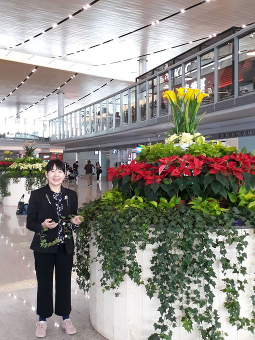 Frau Prof. Dr. Khaing Khaing San, asiatisch aussehende Dame, steht in einer Halle, neben ihr eine große Pflanze mit Efeu in der Hand.