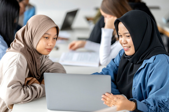 Das Bild zeigt zwei junge Frauen mit Kopftüchern, die gemeinsam an einem Laptop arbeiten. Die Frau auf der linken Seite trägt ein beigefarbenes Kopftuch und einen passenden Pullover, während die Frau auf der rechten Seite ein schwarzes Kopftuch und eine blaue Bluse trägt. Sie lächeln und wirken konzentriert, während sie auf den Bildschirm schauen. Im Hintergrund sind weitere Studierende in einem modernen Lernumfeld zu sehen. Sie sitzen an Tischen mit Laptops und Schreibunterlagen, was auf eine produktive Arbeitsatmosphäre hindeutet. Die Umgebung ist hell und freundlich, mit natürlichem Licht, das durch große Fenster einfällt.