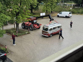 Roter Teleskoplader hebt einen alten weißen und roten VW-Bus in einem gepflasterten Innenhof, zwei Männer beobachten die Szene, im Hintergrund ein weißer Lieferwagen