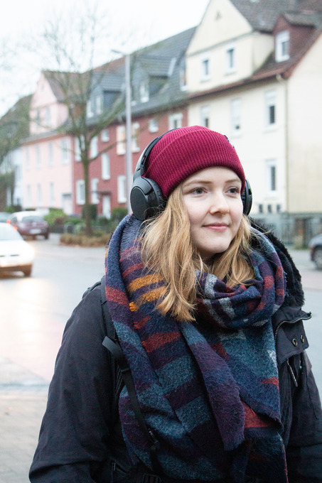 Julia Pohl steht an einer Straße mit schwarzen Kopfhörern auf. Sie hört gespannt den Audiodateien zu.