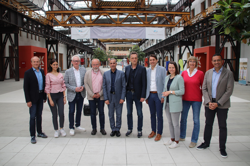 Eine Gruppe von zehn Personen, die in einer geräumigen, industriell gestalteten Halle stehen. Die Halle hat hohe Decken mit sichtbaren Stahlträgern und großen Fenstern, die viel Licht hereinlassen. Ein Banner im Hintergrund trägt die Aufschrift „Herzlich willkommen auf dem Campus Lingen!“, was auf den Standort der Veranstaltung hinweist. Die Personen sind in zwei Reihen aufgestellt und blicken freundlich in die Kamera. Sie sind unterschiedlich gekleidet, von formellen Anzügen bis hin zu Business-Casual-Outfits. Die Umgebung vermittelt einen modernen und industriellen Charme, und die Gruppe scheint für ein formelles Treffen oder einen besonderen Anlass zusammengekommen zu sein. Die Atmosphäre wirkt professionell und einladend.