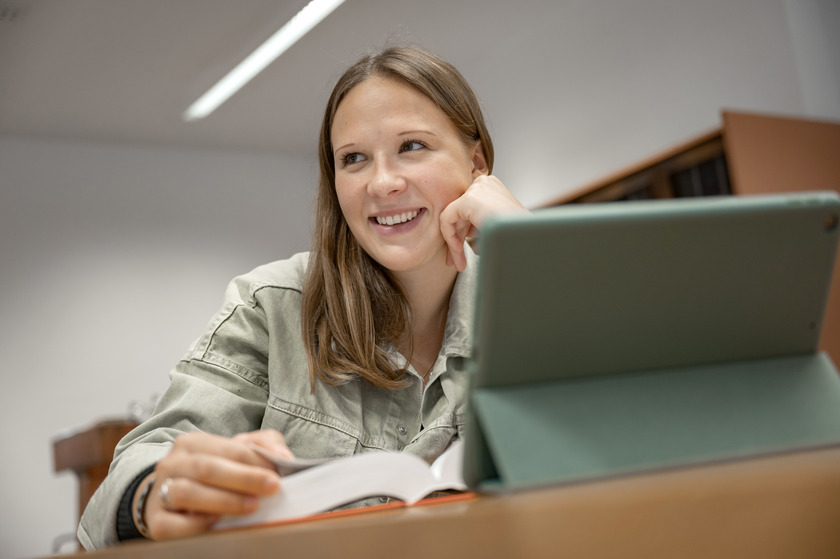 Eine Studentin sitzt mit einem aufgeschlagenen Buch vor ihrem Tablet.