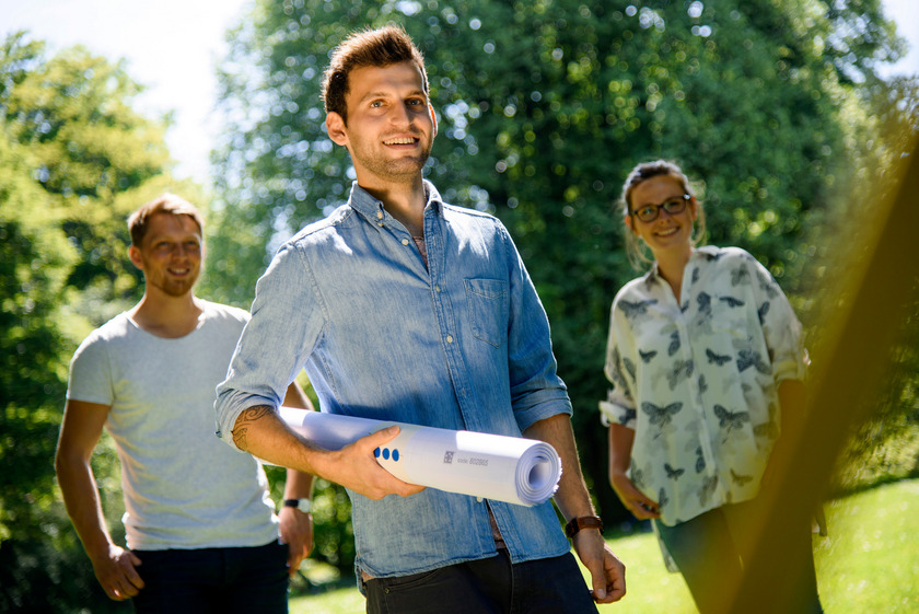 Drei Studierende stehen auf einem grünen Campus. Ein Studierender hält einen zusammengerollten Bauplan unter dem Arm.