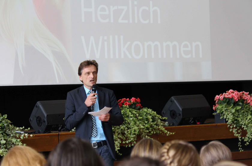 Man sieht einen Mann im Anzug mit Mirko vor einer sitzenden Menge.