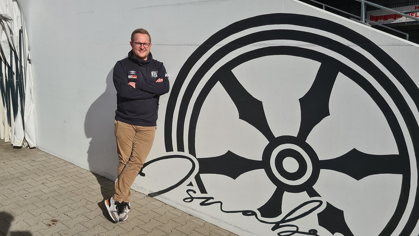 Das Bild zeigt Markus Dörenkämper, der sich an eine Wand im Fußballstadion des VfL Osnabrück lehnt. Er trägt einen schwarzen Vereinspullover, eine beige Jeans und Sneaker. 