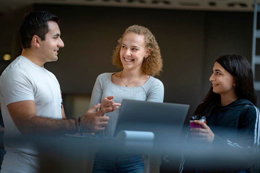 Auf dem Foto sind drei Studierende zu sehen, die vor einem Laptop stehen und sich unterhalten.