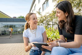 Der Campus Haste lädt zum Lernen im Grünen ein. Zwei Studierende haben ein Tablet in der Hand und lachen sich an.
