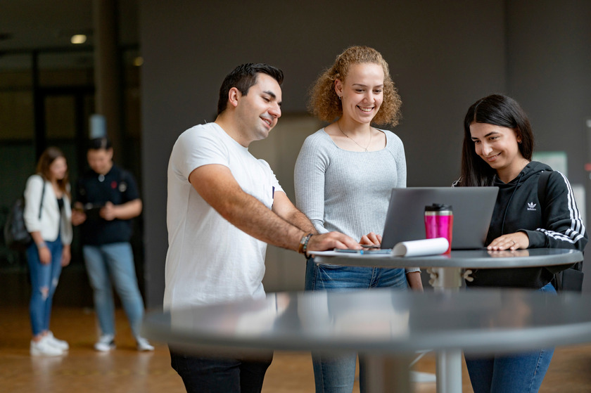 Drei Studierende stehen an einem Stehtisch, unterhalten sich und schauen auf einen Laptop