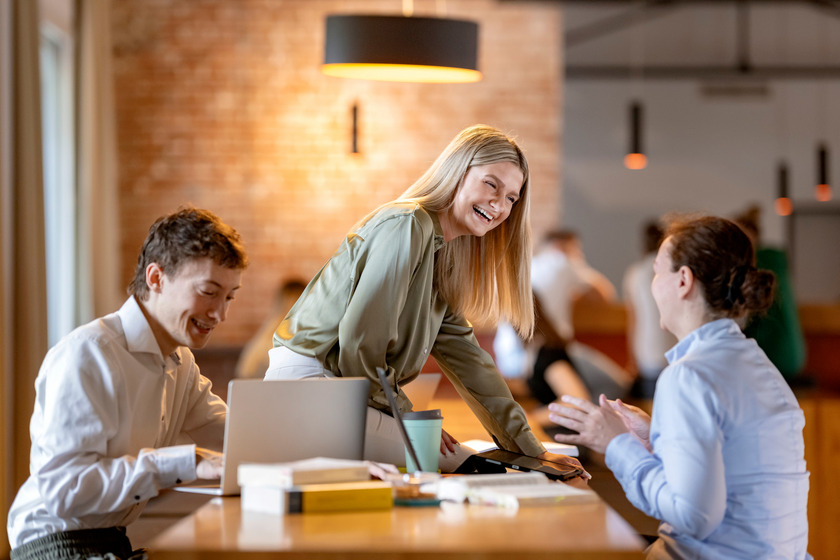Auf dem Foto sind drei Studierende zu sehen. Eine Person arbeitet an einem Laptop und zwei weitere unterhalten sich und lachen dabei.