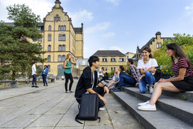 Das Bild zeigt Studierende, die auf einem großen Campus sitzen. Im Hintergrund sieht man schöne, alte Gebäude.