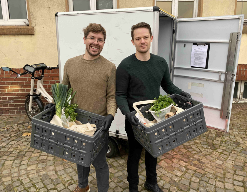 Zwei Männer, die Boxen mit Lebensmitteln in die Kamera halten. Im Hintergrund steht ein Lastenfahrrad.