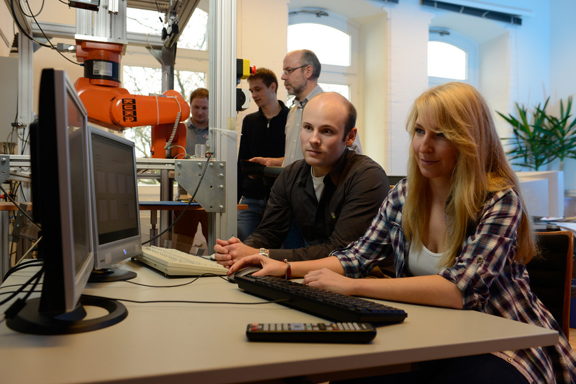 Zwei junge Menschen sitzen vor einem Computer. Im Hintergrund sieht man drei Personen an einer Maschine stehen.
