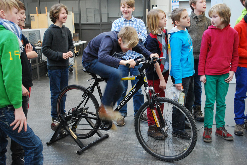 Auf dem Bild sieht man einen Jungen, der sich auf einem Stehfahrrad abstrampelt. Um ihn herum stehen weitere Kinder.