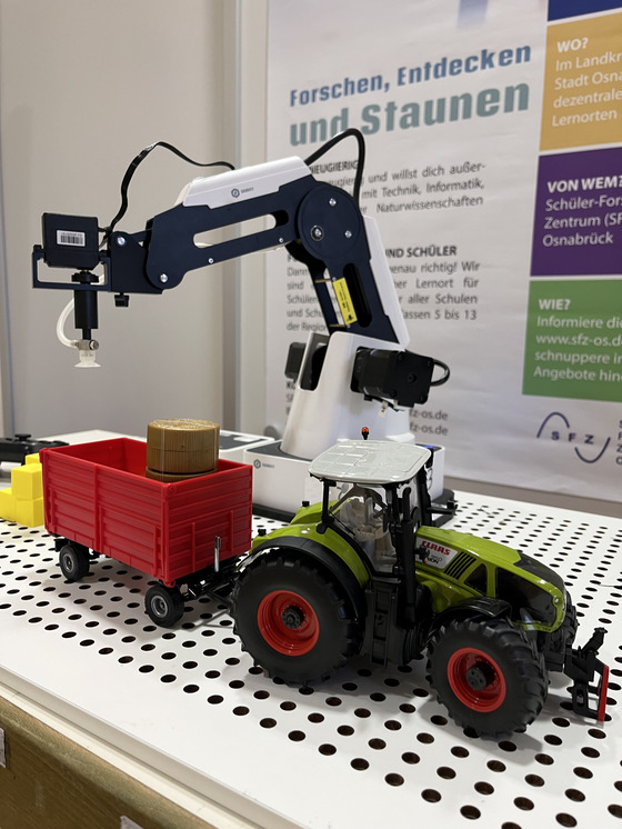 Ein Traktormodell steht vor einem tischgroßen Roboterarm, der den Traktoranhänger mit gelben Plastikwürfeln belädt.