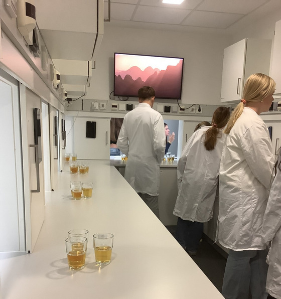 Ein Schüler und zwei Schülerinnen in weißen Laborkitteln stehen in einem Sensoriklabor und testen drei Sorten Apfelsäfte.