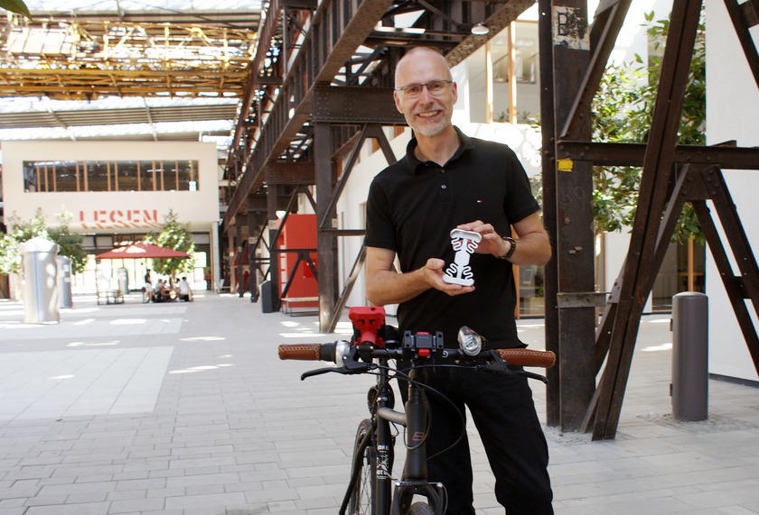 Prof. Dr.-Ing. Jürgen Adamek hält die Halterung in die Kamera. Links neben ihm steht ein Fahrrad.