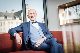 Ein Mann mittleren Alters, ohne Haare mit Brille, sitzt in einem blauen Anzug mit Weste und einer dunkelblauen Fliege auf einem roten Sofa vor einer großen Fensterscheibe. Er lächelt in die Kamera, während sein rechter Arm auf der Sofalehne liegt, der andere auf seinem Schoß.