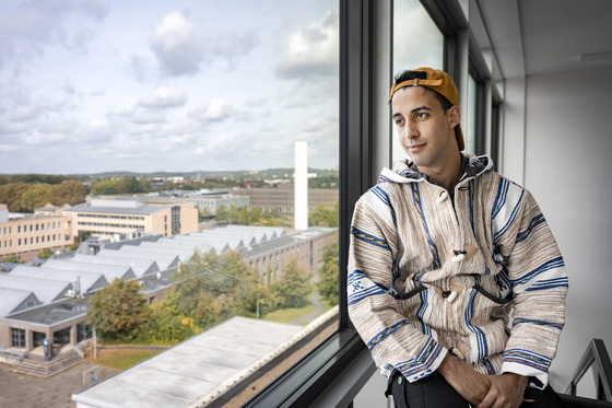 Ein nachdenklicher junger Mann mit olivfarbener Haut und leichtem Lächeln schaut zur Seite und sitzt in einem Innenraum neben einem Fenster, durch das man auf eine städtische Landschaft blickt. Er trägt eine orangefarbene Mütze und ein langärmeliges Oberteil mit ethnischen Mustern in Weiß-, Beige- und Blautönen. Seine Arme sind vor ihm verschränkt, und der Hintergrund bietet einen Blick auf mehrere Gebäude, Bäume und einen bewölkten Himmel.