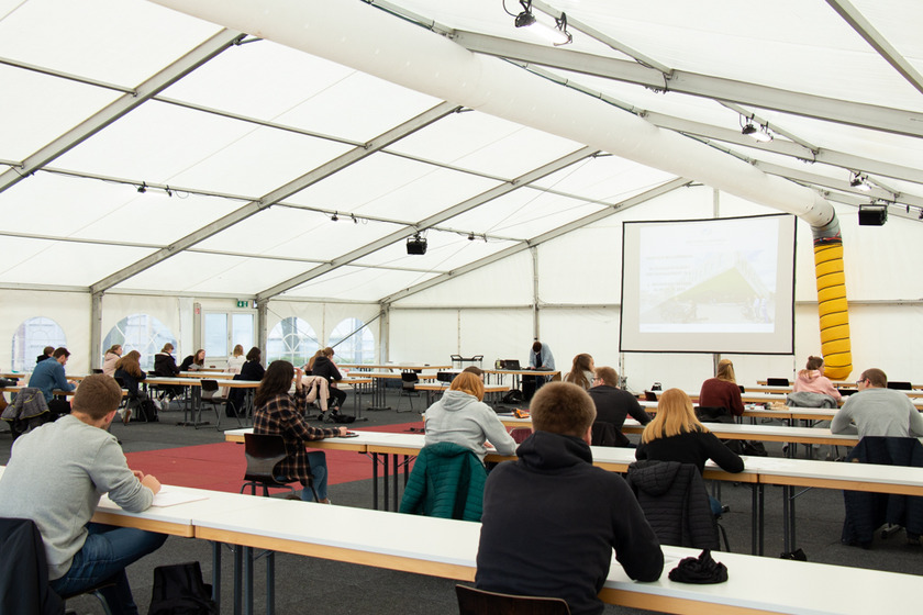 Studierende sitzen in einem Zelt an Tischen und hören den Referierenden gespannt zu. 