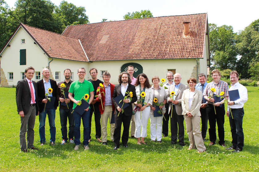 Man sieht eine große Gruppe von Menschen mit Sonnenblumen vor einem Hof stehen.