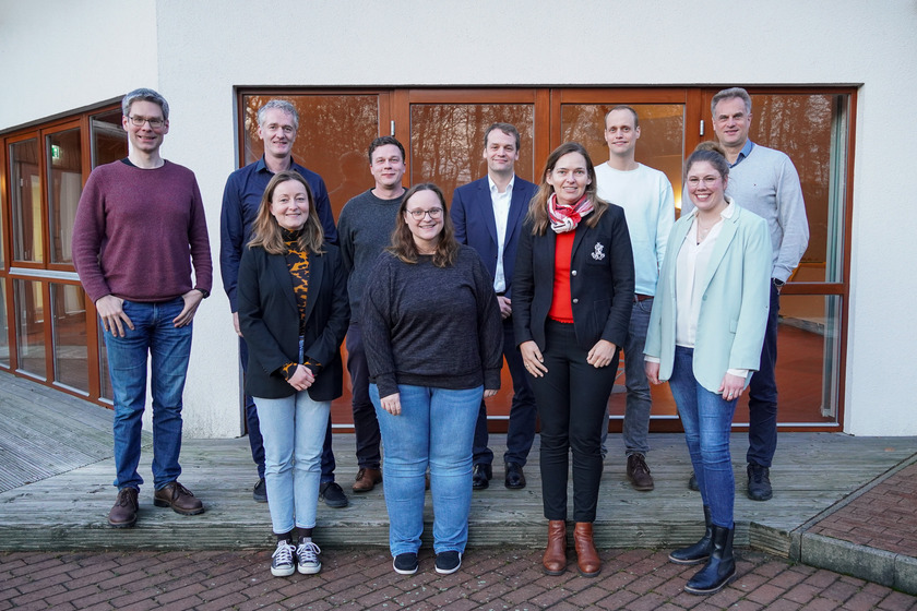 Eine Gruppe aus sechs männlich gelesenen und drei weiblich gelesenen Menschen stehen draußen vor einem großen Fenster. Sie lachen freundlich in die Kamera.