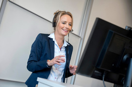 Das Bild zeigt eine Frau mittleren Alters, die mit einem Headset vor einem Laptop steht und redet.