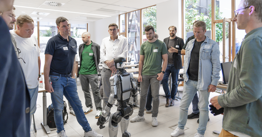 Eine Gruppe Männer stehen im Halbkreis um einen humanoiden Roboter herum. 