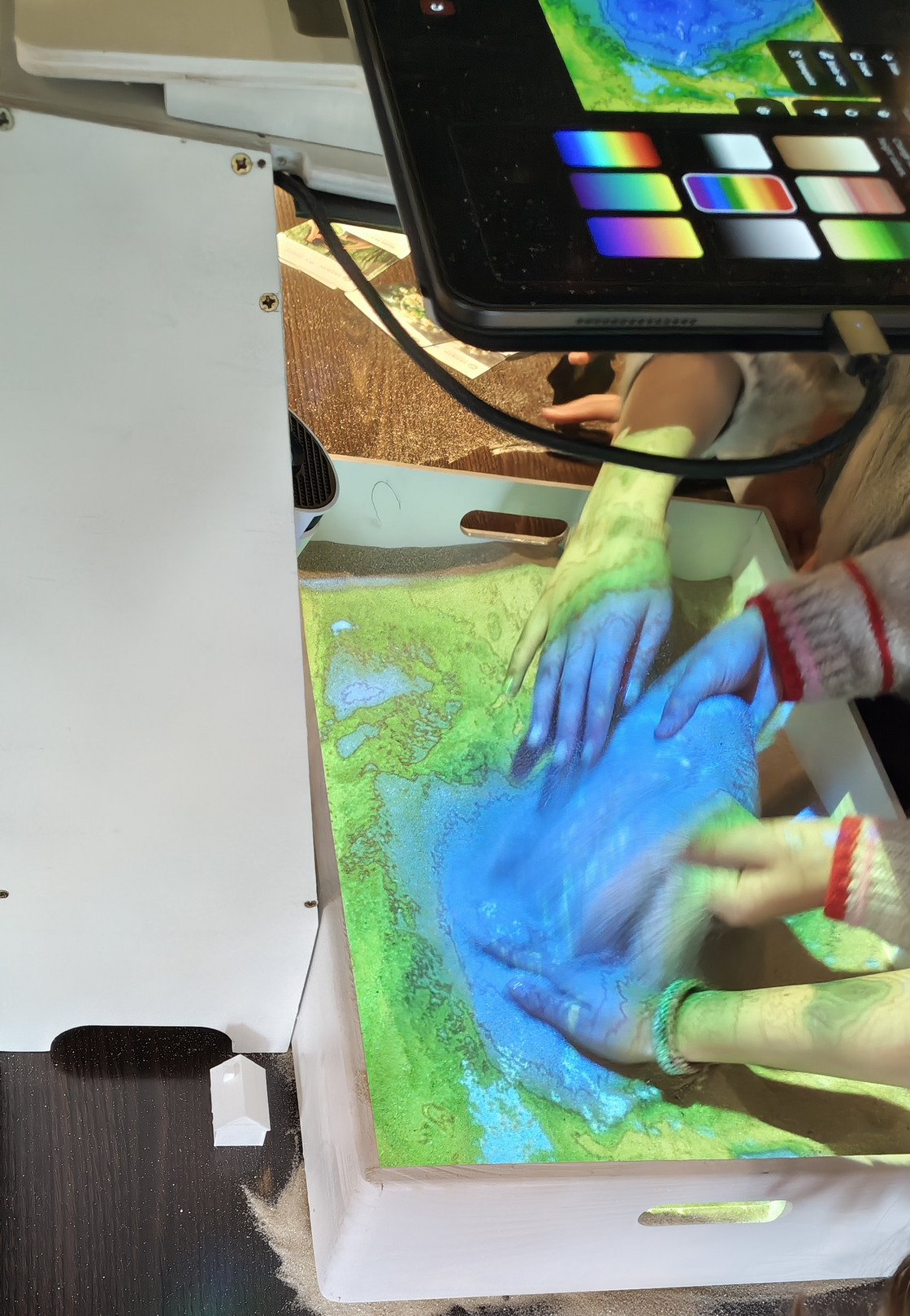 Zu sehen ist eine kleine Version der AR-Sandbox, die von Kindern bedient wird. Im oberen Teil des Bildes ist ein Tablet zu erkennen, über die die Sandbox gesteuert werden kann