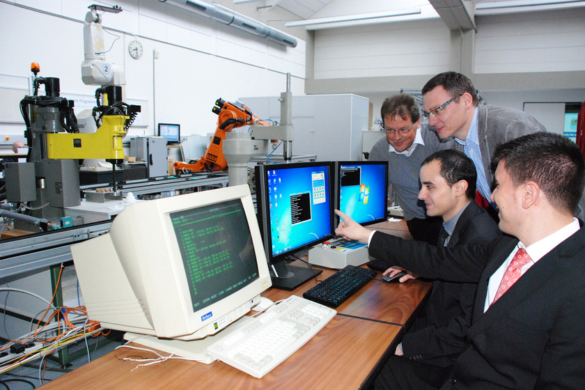 Auf dem Bild sieht man zwei junge Männer in Anzügen vor Computerbildschirmen sitzen. Zwei weitere Männer schauen Ihnen über die Schulter.